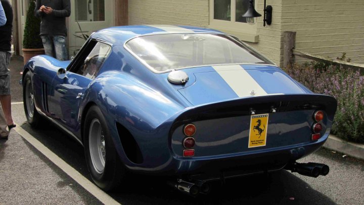 Hare Breakfast Meet - August - Page 1 - Kent & Essex - PistonHeads - The image shows a classic sports car parked by the side of a road. The car is blue with white stripes, adding to its sporty appearance. It features a sleek design with a prominent hood and a small katimus logo on the rear, indicating it's an Italian sports car. The car is parked near a building with another person seen in the background, suggesting an urban setting.