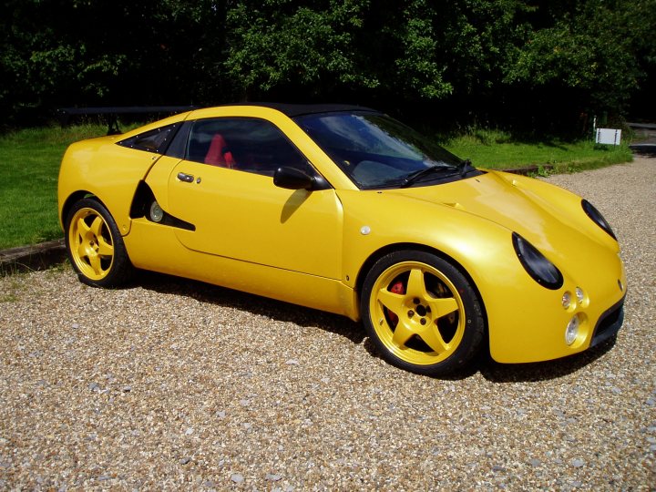 The Best looking Kit Cars - Page 1 - Kit Cars - PistonHeads - The image depicts a single, bright yellow sports car parked on a gravel driveway. It appears to be a two-seater convertible with a sleek, low-profile design, characterized by a long hood and large wheels with visible bolts. The vehicle has a convertible top that is down, revealing a single-piece rear window. It's positioned at an angle to the camera, allowing a clear view of the car's front and side profile. The background features a lush, green landscape, suggesting that the car is parked in an outdoor, possibly rural, setting.