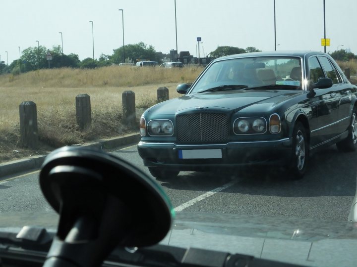 Pair of 'R's - Page 1 - Rolls Royce & Bentley - PistonHeads - The image features a large, black vehicle traveling through an urban setting with a clear sky. The car is seen from a side angle, showing its distinctive grille and front headlights. The vehicle appears to be a modern, high-end car with a large, prominent grille design. In the background, there are several cars parked, possibly in a parking lot, and a street sign can be seen in the distance. The photo is taken through a car window, which suggests the viewer is inside a vehicle.