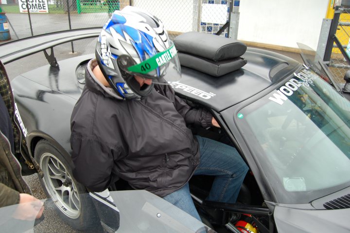 Pistonheads - In the image, a person is standing next to a black sports car. The car features a prominent rear wing and a distinctive "Woods" sticker on the back window. The person is wearing a helmet with a design on it, and appears to be in the process of either examining the car or perhaps preparing for a ride. The car has ghosted hands around it, indicating a level of interaction. The setting seems to be a public outdoor area, possibly a parking lot or a showroom.