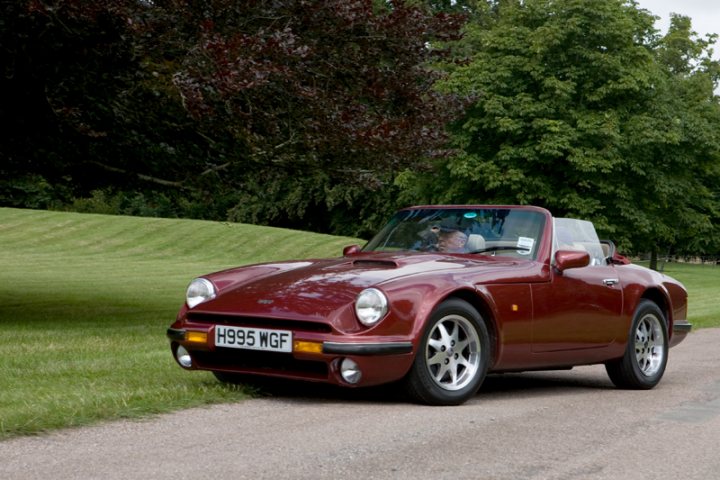 Manor Motors Pistonheads - The image showcases a vintage, red sports car on a tree-lined street. The car is parked on the side of the road, facing towards the left side of the image. The license plate of the car reads "H99 WGF". The backdrop of the scene is a serene suburban setting with green grass and a clear sky around. The overall atmosphere evokes a sense of tranquility, with the car and the surrounding environment creating a harmonious composition.