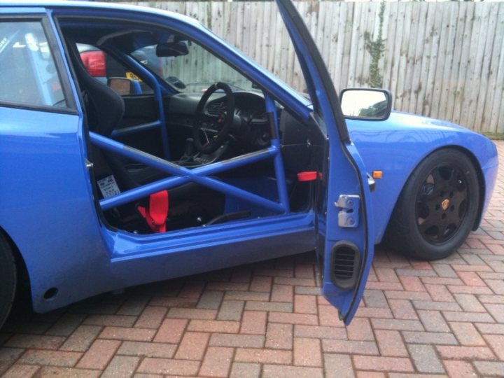 Motorsports Pistonheads Emc - This image captures the driver's side door of a vibrant blue sports car, which is slightly ajar, revealing a red safety strap and the interior darkness. The car is parked on a residence street with a wooden fence in the background, grounded by stone paving. Behind the car, there is a grassy area, possibly a lawn, indicating a suburban setting with a sense of tranquility and privacy. The car's close proximity to the fence suggests a park-like or residential driveway.