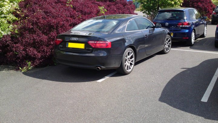 Parking Bad Thread Pistonheads - The image shows two cars parked next to each other in a parking lot. The car in the foreground is a blue Audi, while the car in the background is also blue, but appears to be a different make. Both vehicles are parked within the designated white lines typically used in parking lots to mark parking spaces. The lot itself is paved, with a blacktop visible, and there are red flowers or plants along the edge, suggesting some landscaping efforts within the parking area.