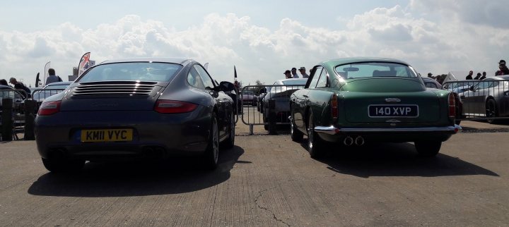 Classics dwarfed by moderns - Page 131 - Classic Cars and Yesterday's Heroes - PistonHeads UK - The image shows a parking lot with two cars. Both cars are sports models, one is black and the other is green. They are parked side by side in a gravel area, facing towards the left of the frame. The background features a cloudy sky and there appears to be an audience watching from the right edge of the image. There's no text visible in this image.