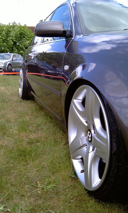 Cars On The Green - Photo Thread - Page 1 - East Anglia - PistonHeads - The image shows a close-up view of the front wheel and tire of a black car. The wheel is silver and appears to be a five-spoke alloy. The car is parked on grass, and there are two other cars visible in the background. The sky in the background is clear with a few clouds. The setting suggests an outdoor event or gathering.