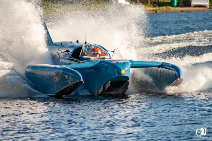 Bluebird K7 Latest - Page 4 - Boats, Planes & Trains - PistonHeads UK - In the image, a blue speed boat is in motion on water, leaving behind a spray of white foam. The boat, with its pointed nose and twin outboard motors, is being driven by a single person who is leaning to the side, indicating high speed or a sharp turn. There are no visible texts or other objects in the image. The background suggests that it could be a lake or river, with calm water and no other boats or people in sight. The photo captures a dynamic moment of hydroplaning, which adds a sense of excitement and motion to the scene.