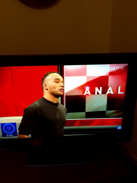 Juvenile things that make you snigger. - Page 163 - The Lounge - PistonHeads - The image is a close-up of a television screen displaying a man in a black T-shirt standing in front of a checkered background. The screen is showing an advertisement that features a logo of an adult brand. The logo consists of the word "ANAL" with stylized letters and checkered patterns. Above the logo, there is text that indicates the digital channel, such as "E4". The time "01:00" is displayed in the upper left corner of the screen. The photograph captures this snippet of television content with a focus on the advertisement.