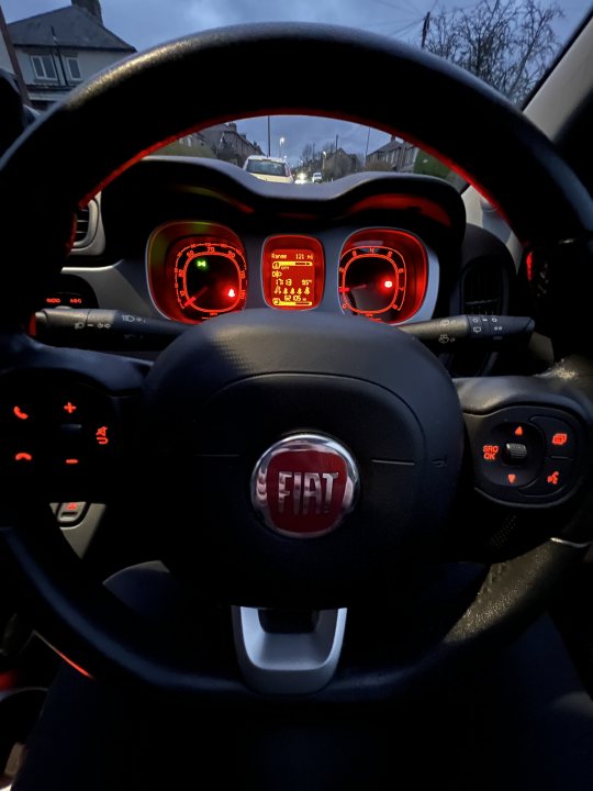 Pistonheads - The image shows an interior view of a car from the driver's perspective. The steering wheel is in the foreground, featuring a red and black design with several buttons and gauges. Behind the wheel, there are two dials or gauges, likely for speed and revolutions per minute. The car has a black leather interior, with red accents visible on the door panels and gear lever. The dashboard is illuminated by the instrument lights, emphasizing the red and black color scheme of the vehicle. The car appears to be parked indoors, as there's a glimpse of what looks like a domestic setting through the windshield.