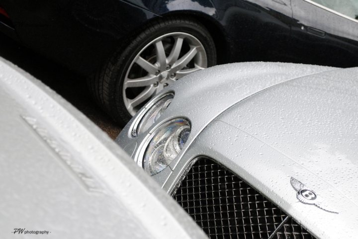 I’ve put my football boots on & bought a Bentley GT Speed - Page 1 - Rolls Royce & Bentley - PistonHeads - The image captures an intimate battle between two luxury vehicles, both coated in a glossy rain, reflecting the light in their silver and black exteriors. The cars are parked on a street, their stance facing the camera. The car in the foreground is a sleek silver vehicle, while the one in the background is a classic black sports car. The wet surfaces add a certain sheen to their appearance, accentuating their polished exteriors. The contours of their bodies and the lower halves of their grills can be seen, filling the frame with their modern designs.