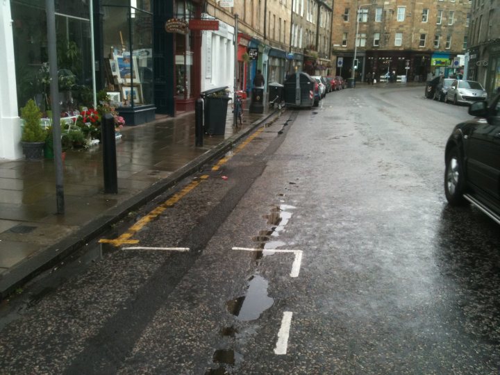 Single yellow line - Fine but no sign - Page 1 - Roads - PistonHeads - The image depicts a rainy day on a city street lined with shops. The street appears to be slick from the rain, as indicated by the reflections on the wet pavement. Parked cars are visible along the side of the road, and people can be seen walking on the sidewalk, taking shelter from the rain. The shops have displays of flowers and plants at their entrances, adding a touch of nature to the urban setting. Overall, the image captures a typical rainy day in a bustling city.