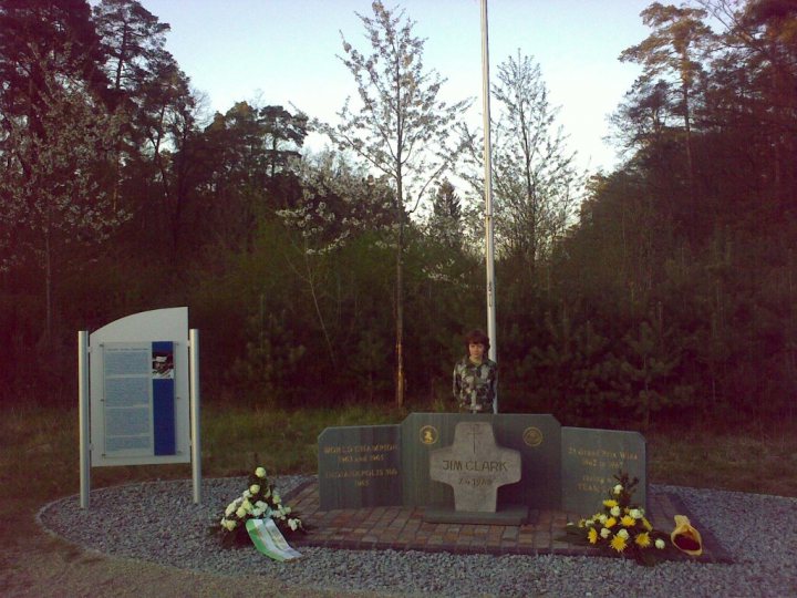 Jim Hockenheimring Pistonheads Clark - In the serene setting of a memorial, a young boy, standing to the left of the scene, faces a monument. The monument, constructed from stone, rests on a gravel base. The memorial is adorned with flowers, including a white cross placed prominently above a green plaque. The backdrop is a tranquil forest, adding a sense of peace to the poignant scene.