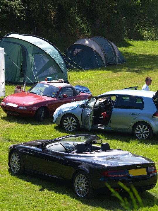 Stickering up for 2016 - Page 1 - Le Mans - PistonHeads - The image showcases various vehicles parked on a lush green field, with a vibrant mix of cars and a Volkswagen camper. The cars vary in color from metallic red to sleek black, and a car with a large backward-facing window catches the eye. Behind the vehicles, a camping scene unfolds with multiple tents, including a sizeable hexagonal one. A person can be seen at a distance, adding a sense of scale to the scene. The setting appears to be a casual, outdoor gathering, possibly a camping trip or a small car show.