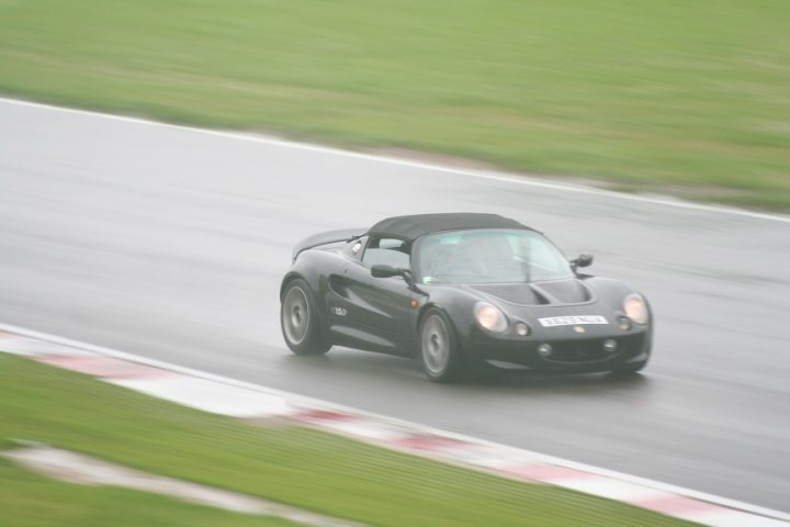 Elise Sport 160 Honda Conversion - Page 1 - Elise/Exige/Europa/340R - PistonHeads - This image captures a scene on a racetrack, where a black convertible sports car is in motion around a bend. The car's top is down, suggesting a good weather condition, and it's driving on the inside lane. The image is slightly out of focus, adding a sense of speed and movement.