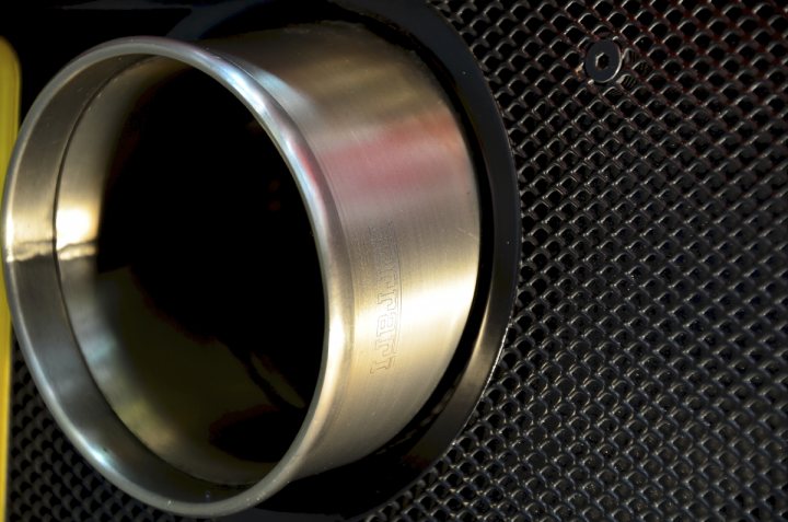 A close up of a mirror on a wall - Pistonheads - The image captures a close-up view of a shiny, cylindrical object, which appears to be an exhaust pipe or an engine part. The object is plated in a metallic gold color, reflecting some red hues, suggesting a surface possibly treated or coated in a specific material. Positioned behind this metallic piece is a perforated black or dark grey panel, giving a contrast between the two textures and colors. A small black screw or bolt is visible in that black surface, adding another layer of detail to the composition. The photograph conveys a perspective that emphasizes the golden object on the left while the perforated surface creates an interesting backdrop.