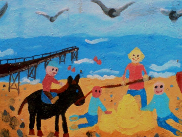 Spring Air Restoration Pistonheads - The image shows a colorful mural painting on a wall of a scenic beachside. It features three children, a dog riding on a cart, and a character combining the features of a human and a dog. The painting is composed with broad brushstrokes, achieving a lively and cheerful atmosphere. The style appears to be folk art, characterized by its simplicity and vibrant use of color.