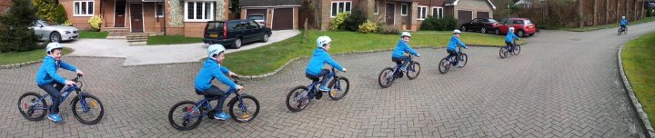 A group of people riding bikes down a street - Pistonheads - The image shows a row of people riding bicycles on a cobblestone street. They are wearing matching blue jackets, indicating they might be part of a group or event. At the end of the line, a person walks alongside the bicyclists. The setting appears to be a residential area with houses on either side of the road. The overall atmosphere of the image is recreational and cycling-focused.