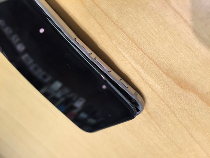 A cell phone sitting on top of a wooden table - Pistonheads - The image captures a close-up view of a smartphone, which appears to be an HTC phone with a model number visible. The phone is resting on a wooden surface, the grain of the wood running vertically and horizontally. A noticeable scratch on the top left corner of the phone's surface is illuminated by the phone's display. The screen is turned off, with the circular flashlight at the top.