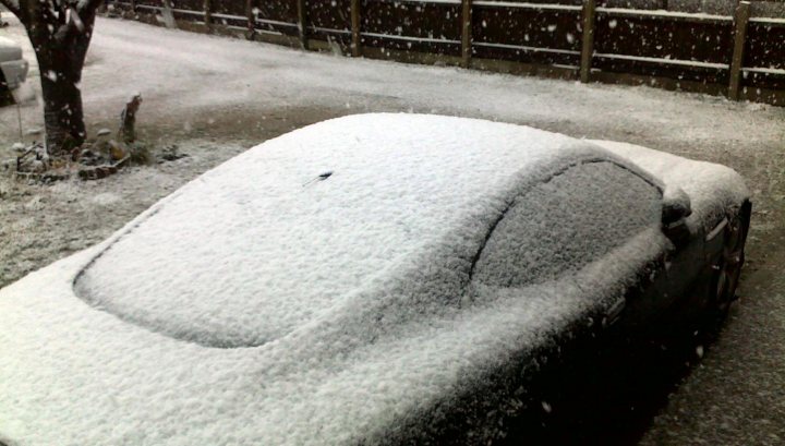 Any snow? - Page 1 - North West - PistonHeads - The image shows a scene of a car covered in snow. The car is an all-black model, characterized by a glossy finish. Its rear window, roof, and hood are heavily dusted with snow, indicating that it has been parked outdoors during a snowstorm. The vehicle appears to be the main subject of the photograph. The photo is taken from a slightly elevated angle, possibly from a second story window or similarly high vantage point. The background includes a portion of a fence, suggesting that the car is parked in a residential area. The photo is slightly blurred, further emphasizing the inclement weather conditions during which it was taken.