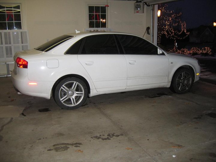 The image shows a white car parked inside a garage. The car appears to be an older model with a formal style, characterized by the presence of a sunroof. The garage has lights on, illuminating the car and the immediate surroundings. The car has black wheels and is positioned at the side, with the car's rear wheel visible in the foreground of the image. There is no text or person in the picture, and the garage door can be slightly seen in the background, indicating the car is parked close to it.