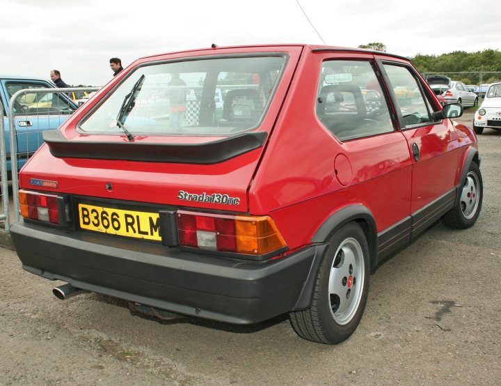 Your favourite car from the year you were born in... - Page 12 - General Gassing - PistonHeads - The image showcases a British-style red car with a black roof rack, parked on a gravel surface. The car has a license plate that reads "B36 RLM". The backdrop is a parking lot with other vehicles and some people in the distance, suggesting an outdoor car event. The car's design and color scheme are striking, making it the focal point of the photo. Despite the mechanical subject, the overall vibe of the image is casual and relaxed, likely capturing a leisurely day out at the show.