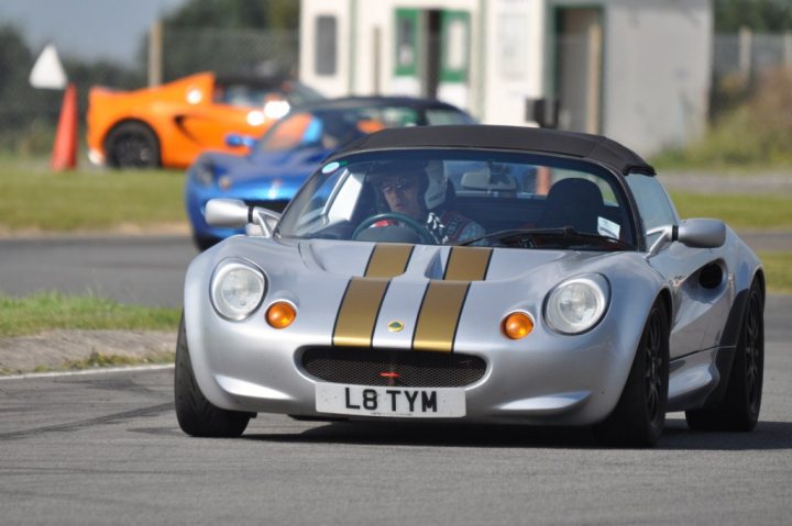 Can you make graphite grey elise stand out more? - Page 1 - Elise/Exige/Europa/340R - PistonHeads - The image shows a silver sports car with a distinctive black and gold stripe across its body, driving on a road. One person, potentially the driver, is visible through the car's window. Behind the sports car, there's another vehicle, and a grassy area with trees is visible in the background. The sky is overcast, indicating a cloudy day.