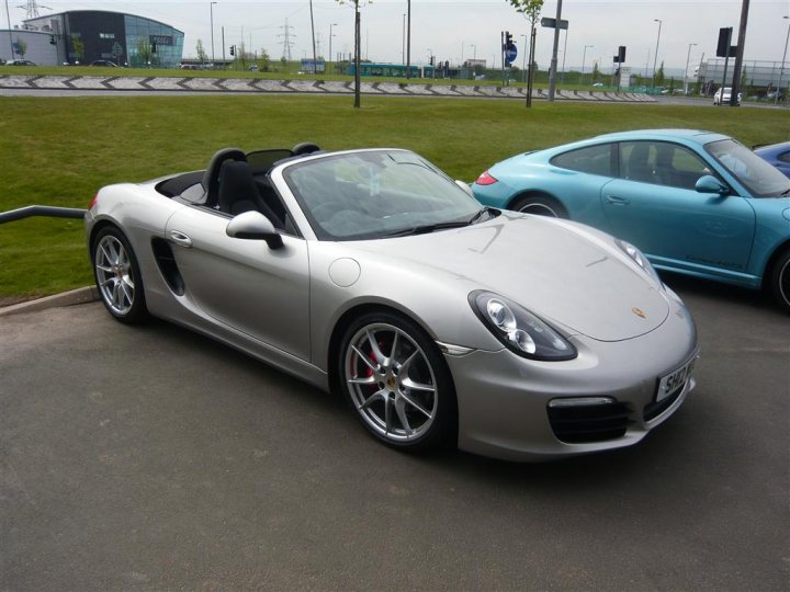 NEW 981 BOXSTER OWNERS - PROSPECTIVE PURCHASERS FORUM - Page 16 - Porsche General - PistonHeads - This image captures a scene on a sunny day in a parking lot. In the foreground, a silver convertible Porsche sports car is parked. It has a three-pointed star on the front grille and a side exhaust pipe. The car's doors and the top are open, revealing the interior and the driver's seat.

Behind the Porsche, there is a blue sports car. It has a distinctive round headlight design and is also parked with its doors open. In the far background, parallel to the cars and across the road, there are commercial buildings with streetlights lining the roadside.