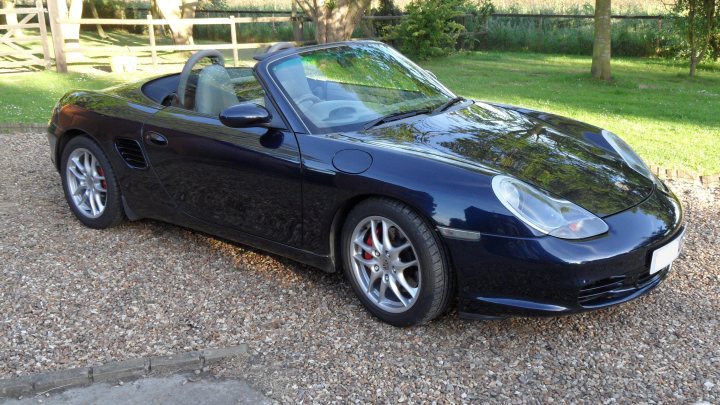 986 Boxster S Buying & Spec Advice - Page 1 - Boxster/Cayman - PistonHeads - The image shows a black convertible car parked on a gravel surface. The car is a two-door convertible, with the top down, hinting at the courteous weather. It is angled towards the left side of the frame, giving a clear view of its front end and the flagrant red Rotors on the wheels, which add a splash of color against the car's dark paint. In the background, the lush greenery of a field or garden provides a serene and natural backdrop to this grand vehicle. The overall image conveys a sense of luxury and enjoyment of outdoor drives.