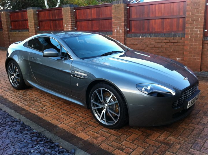 Looking for  pics of 2010 or 2011 Hammerhead V8? - Page 1 - Aston Martin - PistonHeads - This image features a gray Aston Martin sports car parked on a brick-paved driveway next to a wooden fence. The car appears to be in excellent condition with a sleek design and shiny chrome accents. It's positioned at an angle to the viewer, allowing a clear view of its front wheel and side profile. The surroundings suggest a private residential area.