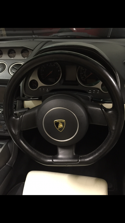 New steering wheel  - Page 1 - Gallardo/Huracan - PistonHeads - This image shows an interior view of a luxury car, centered around the driver's side seat and steering wheel. The steering wheel is large and finished in black leather, onto which a gold or yellow logo is prominently placed in the center. The dashboard has multiple gauges and appears to be a digital or infotainment display. There's a partial view of a door with a door handle visible. The car is bathed in a soft light that casts gentle shadows, indicating indoor lighting.