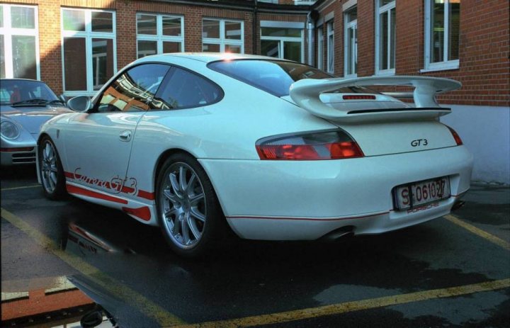 Prospective 981 GT4 Owners Discussion Forum. - Page 71 - Porsche General - PistonHeads - The image depicts a white Porsche 911 GT2 RS parked by the side of the road. The car is facing the viewer and is positioned in front of a building with a brick exterior. The design of the Porsche sports car is reflected in a wet puddle on the road, adding a unique perspective to the image. The car appears to be in good condition, suggesting it might be well-maintained. The area around the car seems quiet and undisturbed.