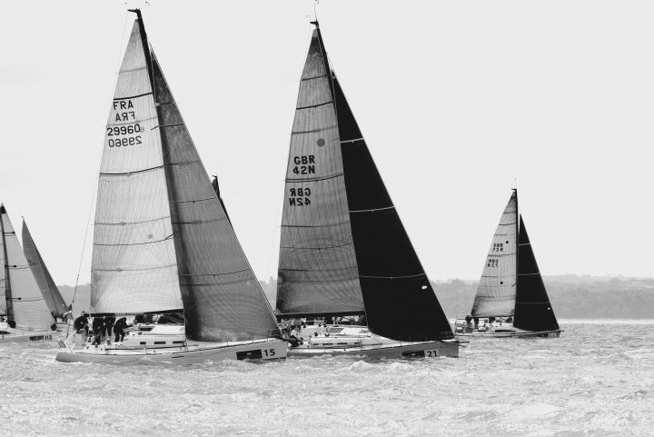 FAO: Folks around the Portsmouth areas with exotic cars - Page 2 - South Coast - PistonHeads - This black and white photo captures the essence of a sailing boat race. The lead boat is prominently featured in the foreground, with several other boats trailing closely behind in what appears to be a challenging regatta on the ocean. Each sail is adorned with wording, likely indicating the boat names or identification numbers. The rings made visible by the water's surface radiate, giving a sense of the boats' locations. The overall action suggests a competitive atmosphere, with each boat jockeying for position.