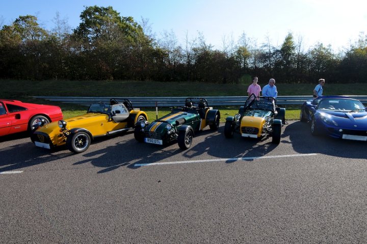 Pistonheads - The image displays a line of seven classic sports cars parked next to each other on a paved area. The cars vary in their vintage design and come in an assortment of colors, including blues, reds, and yellows. The vehicles are parallel parked, with their hoods facing the same direction. In the background, there are some people and trees, which adds a natural feel to the scene. The photo captures the beauty of the cars as they are taken out for an event or gathering.