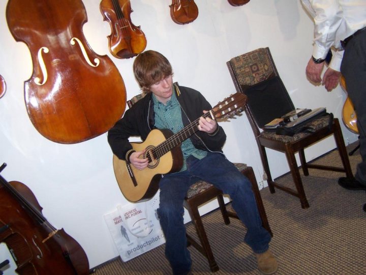 A group of people sitting around a table - The image captures a young man deeply engaged in playing a guitar with a confident posture. He is seated on a chair in a room filled with a curated collection of musical instruments. The room also contains a few other chairs, a couch, and a laptop on a table. The young man is wearing a green shirt, and a second individual is seen in the background, adding a sense of the space. The overall ambiance suggests a relaxed, creative environment, possibly a home or a local music venue.