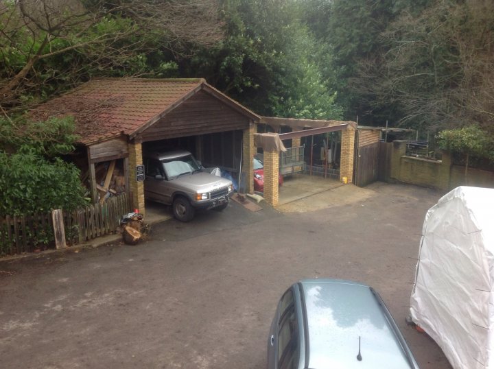 And so it begins.................... - Page 7 - Chimaera - PistonHeads - The image shows a covered parking space outside a storage or workshop building. Inside the covered area, a silver truck is parked. Another silver vehicle is parked in front of the covered area. The ground appears wet, suggesting recent rain. The setting is tucked away in a wooded environment, and a sign is visible in the distance.