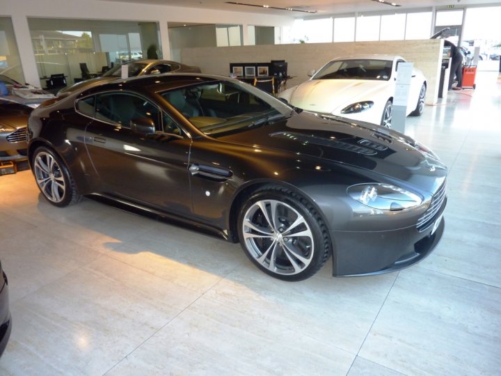 Pistonheads - The image presents a dealership display room, where a dark-colored sports car with a sleek design and chrome rims is prominently featured. The car is positioned on a reflective floor, adding to its polished appearance. In the background, other vehicles can be observed, suggesting a variety of makes and models on display. The dealership seems to be well-lit, and the overall atmosphere is that of a sophisticated, high-end sales environment.