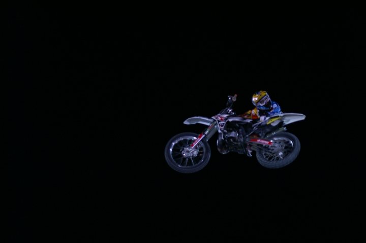 Red Bull Pistonheads Fighters - The image features a dynamic scene of a motocross rider launching off a jump on a dirt bike. The rider, depicted in full racing gear, is positioned at the top of a sandy hill, his arms wide for balance as he prepares to land. The motorcycle has a prominent front wheel, indicating mid-flight, and is angled toward the left of the image, adding a sense of speed and motion. The dark background accentuates the rider and bike, creating a dramatic contrast between the high-energy subject and the stillness of its environment.