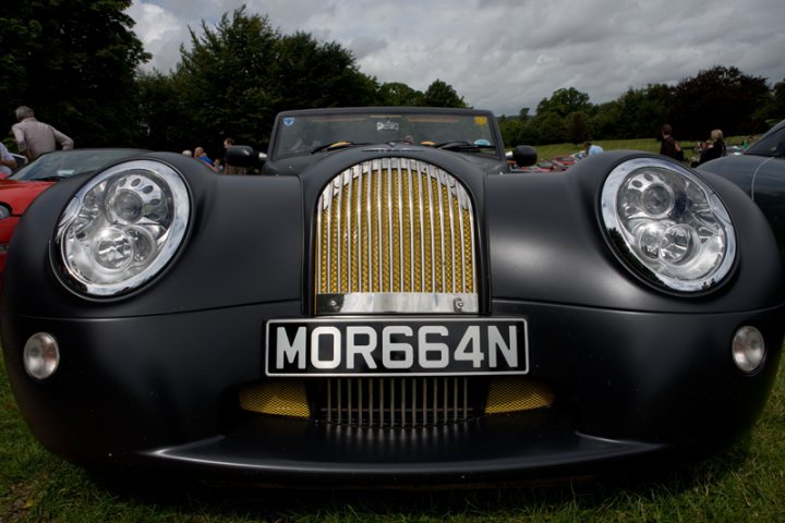 Manor Motors Pistonheads - This is a photograph of an old-fashioned, black car that has a classic design, featuring a prominent front grille and round headlights. The vehicle has a distinctive license plate that reads "MORE 664N" in capital letters. The car is the focal point of the image, parked on a grassy field where other cars are visible in the background, suggesting the car may be at a display or other gathering. The sky overhead is cloudy, contributing to the vintage aesthetic of the photograph.