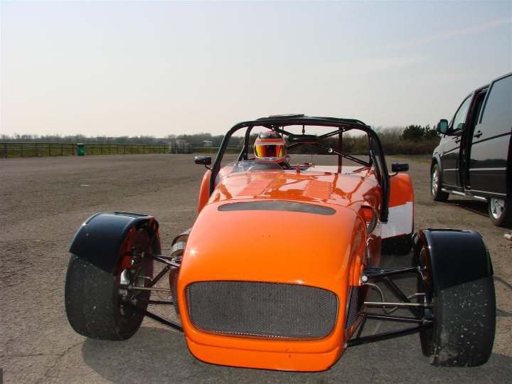 Race Mechanics Pistonheads - In the image, there is a striking orange race car parked on a slightly textured concrete surface. The car has two tires on the side, and a visor is worn on the front. The background shows a clear sky, and there is a figure driving beside the car, adding a dynamic element to the otherwise static scene. The setting suggests a track, and the image conveys a sense of motion and speed, potentially indicating an event or competition.