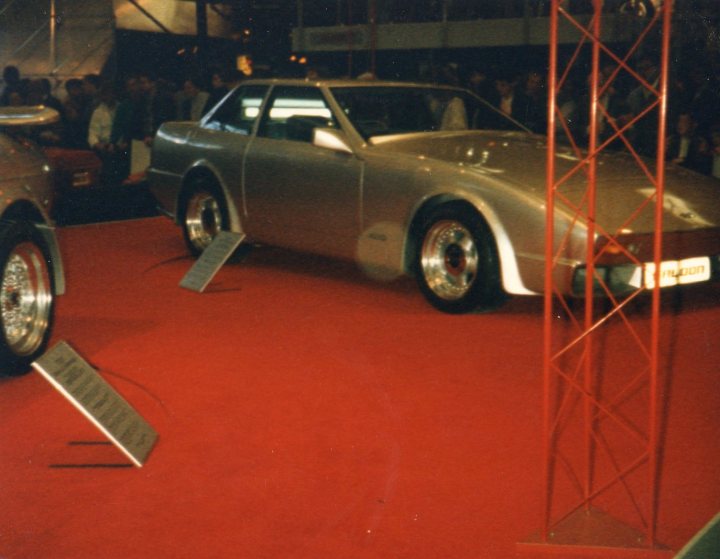 Old Wedge Photo - Page 1 - Wedges - PistonHeads - The image depicts a vintage sports car displayed on a red carpet in what appears to be an auction setting. The car is a classic European two-door model, with a distinctive curved shape, chrome detailing, and black side mirrors. It is parked on a platform with two ramps, likely to facilitate its transport. In the background, there are additional people and cars, suggesting that this is part of a larger event or exhibit.