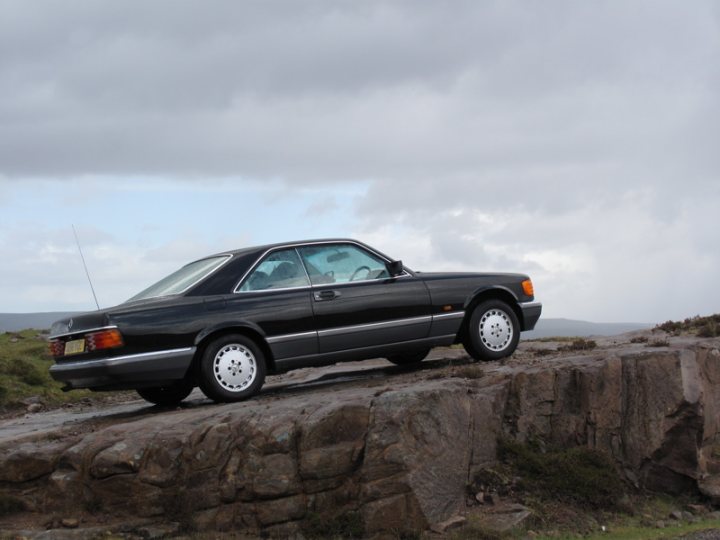 Show us your Mercedes! - Page 78 - Mercedes - PistonHeads - The image shows an old, black car parked at the edge of a grassy cliff with a rocky wall. The setting is overcast, and the car appears to be a European model with a sunroof, illuminated headlights, and distinctive rims on the wheels. The rocks, grass, and the contrast between the man-made luxury of the vehicle and the natural ruggedness create a striking juxtaposition.