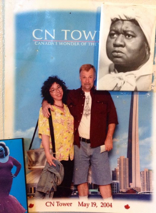 This image appears to be a postcard or a photograph with embellishments. It features a couple in the foreground, smiling and posing together, with a red-haired man in a maroon shirt and a woman wearing a yellow blouse. Behind them is a blue sky and to the right, a prominent skyscraper silhouette and text that mentions "CNN Tower" and "MAY 19, 2004". On the left edge of the image, there's a smaller photo of a person from the Civil Rights era. The overall theme and style of the image suggest it's from a past event, possibly related to television or journalism, celebrating the visit of a famous individual to the locale.