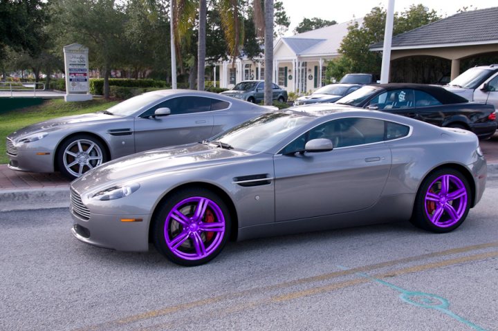 Pistonheads - In the image, a parking lot is filled with several cars, including a pair of silver Aston Martins and a purple and black sports car. The cars are parked along the curb with their fronts facing the camera, showcasing their sleek designs. The surroundings include trees, a lamp post, and a building with a green roof, providing a contrasting backdrop to the shiny, metallic bodies of the sports cars. The photo appears to be taken during daytime under clear skies.