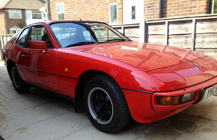 Any good with stereos? - Page 1 - Home Mechanics - PistonHeads - The image shows a classic red sports car parked in front of a building with a red brick exterior. The car is positioned at a slight angle to the camera, allowing a clear view of its front end and driver's side profile. It is a convertible model with a black soft top roof that is currently closed. The vehicle appears to be in good condition, with shiny paintwork and well-maintained road wheels. There are no visible texts on the car or in the immediate vicinity.