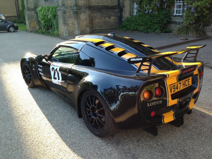 A red car is parked on the side of the road - Pistonheads - An exotic sports car is parked on a driveway. The car's body is predominantly black, adorned with striking yellow and black patterns. A large rear wing, presumably for increased downforce, adds continuity to the vehicle's design. The license plate identifies a valid registration with the tag "91FCE". The surrounding environment is simple yet elegant, consisting of a road, buildings, and greenery, providing a serene backdrop to the car's sleek profile.