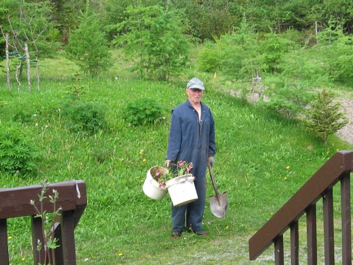 Garden Siton Pistonheads Justify Big Lawnmower - The image shows a person standing in the foreground, holding two white pails in their hands. The person is wearing a cap, a dark jacket, and dark pants, giving the impression of casual outdoor attire. Behind the person, there is a small, neatly trimmed green lawn, and beyond the lawn, there is a path leading into a forested area. To the right of the person, there is a set of wooden stairs leading down into the greenery.