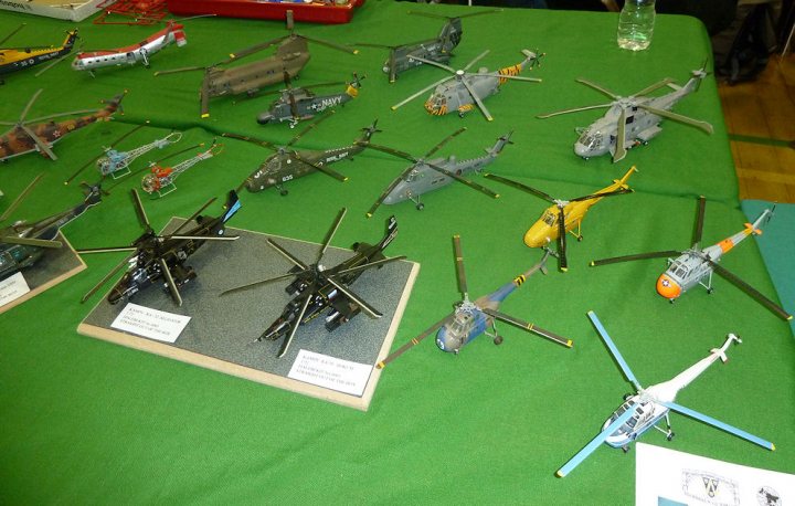 Pistonheads Crewe Model Feb - The image presents a lively scene of a display table exhibiting a variety of toy helicopters. The table, covered with green felt, is a bustle of aerial joy, with twelve of these flying machines arranged in rows. Each helicopter is uniquely colored and designed, their rotors poised for action, adding a touch of excitement to the scene. Above the table, two of the toy helicopters are suspended in the air, almost as if they are in the midst of flight, creating a sense of movement in what is otherwise a stationary display. The helicopters are meticulously displayed, with each type clearly marked, inviting viewers to appreciate the intricate details and craftsmanship of these miniature choppers. The labeling them is cleverly placed in front of each figurine, providing a concise guide for visitors interested in learning more about this delightful array of toy helicopters.