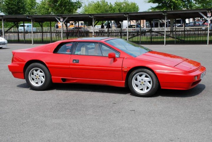 Sunday Pistonheads Supercar - The image displays a bright red sports car with a sleek design, parked on what appears to be an open road or parking lot. The car is viewed from the side, presenting a profile view that showcases its aerodynamic shape, side mirrors, and one of its doors slightly open. In the background, there is a structure resembling a covered garage or storage area with metal frames. There are also several other cars and a truck in the distance, possibly suggesting a parking area or roadway alongside a building or commercial area. The setting seems to be an outdoor location under clear skies.