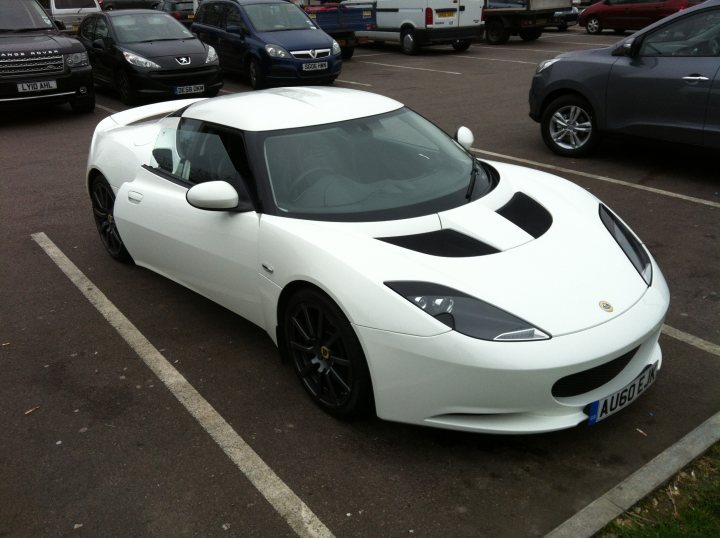 I got one! - Page 2 - Evora - PistonHeads - The image features a white sports car parked in a parking lot. The car is positioned at an angle, with the side view showing a silver door handle. Surrounding the car, there are other vehicles parked in different orientations. The background shows a partially visible white building. The scene appears to be a typical day with daylight illuminating the environment.