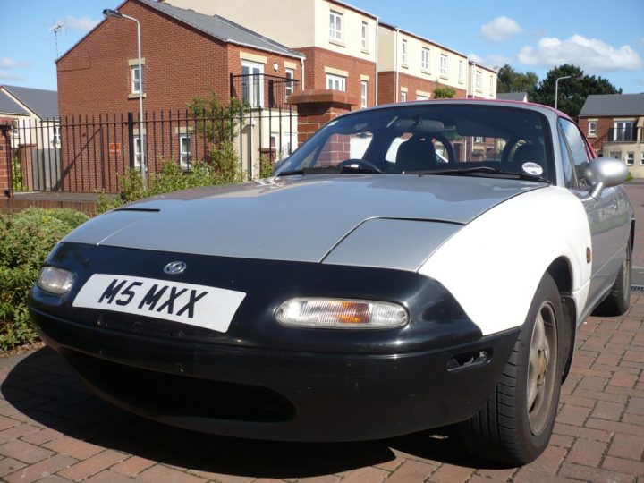 Pistonheads Booked Bodyshop - The image showcases a sleek, white and gray sports car that dominates most of the frame. The car is parked on the side of a street lined with brick paving stones and lush green shrubs. In the backdrop, there's a row of residential houses painted in a mix of beige and red tones. The clear blue sky suggests a sunny day adding to the overall pleasantness of the scene.