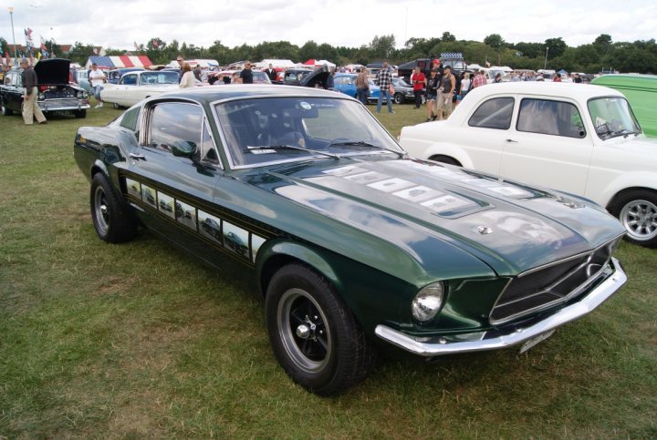 Anyone going to the NASC 40th Nats at Trinity Park? - Page 1 - East Anglia - PistonHeads - The image depicts a vintage green Mustang parked at a car show. The car is positioned prominently in the frame, indicating its importance. It has a sporty design, including a long hood and short rear deck, which is characteristic of classic Mustangs. The Mayhem design on the side implies that the car owner has a penchant for personalizing and tuning their vehicle, a common hobby in the car show circuit. In the background, you can see a multitude of other cars and people, indicating that the Mustang is part of a larger gathering.